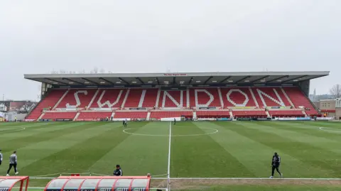 A football stadium stand with the words "Swindon" written in white letters against a red background. Stadium lights and and other buildings can be seen in the background.