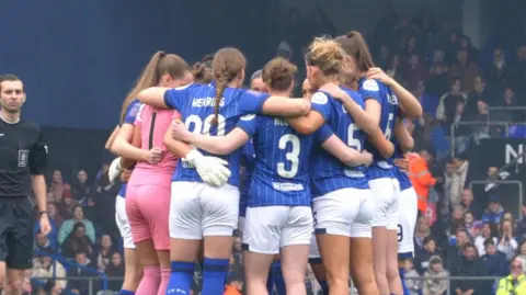 Ipswich Town Women are pictured in a huddle during their Portman Road match. A referee in a black stands nearby while the crowd behind them watches on.