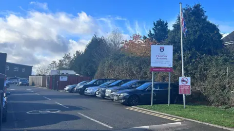 Kate Bradbrook/BBC A photo of the car park outside the school and its sign, next to a flagpole with the British flag on. The edge of the school building can be seen in the photo.