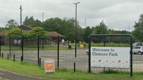 Signage saying 'Welcome to Elemore Park' sits before a fenced off car park. A large patch of green space and buildings are visible beyond the car park.