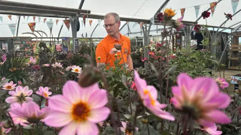 Oliver Whitfield-Miocic/BBC A man in a bright orange shirt is standing inside a garden centre. He is surrounded by pink flowers.