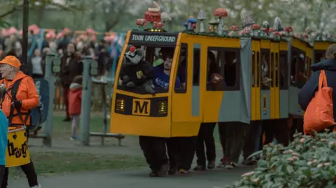 Von Fox Promotions A large puppet of the Tyne and Wear Metro yellow train which is made out of cardboard and has multiple people standing inside of it to make it move. There are various toadstools on the roof of the train and a badger puppet being held up by the person at the front. The destination on the front of the train reads Toon Underground. They are in a park with people around them.