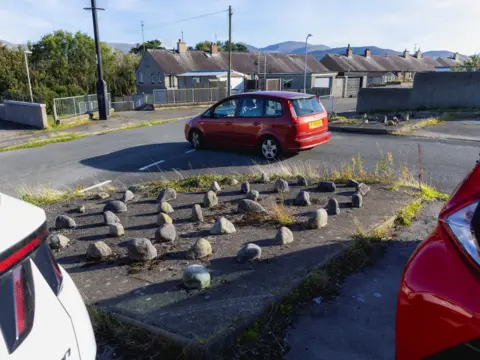 David Hurn A red car is pulling out onto a road at a junction with a row of garages beyond. Behind it is a square of concrete with big stones arranged on top of it, and a glimpse of two cars, one red, one white, parked behind.