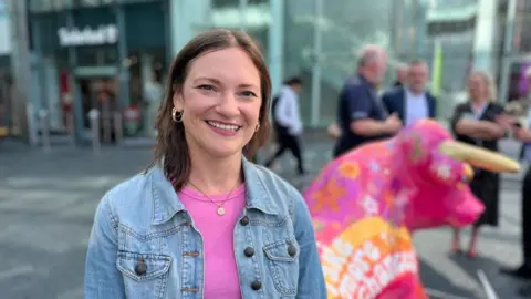 A woman in a denim jacket and wearing a necklace and earrings is smiling looking at the camera. Her mainly pink bull is blurred in the background.