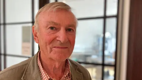 A man is looking into the camera. He's wearing a beige jacket and a checked shirt. There are large windows behind him.