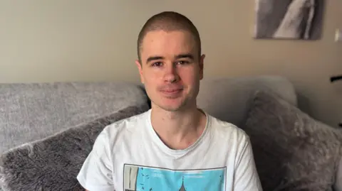 Liam Virgo, wearing a white t-shirt and sitting on a grey sofa.