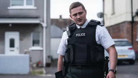 BBC/Long Story TV/Peter Marley Finnian Garbutt, a man with dark hair, wearing a white shirt with black tie and a black support vest with the words police on it. He is wearing a black watch on his left wrist and there is a row of white houses in the background.
