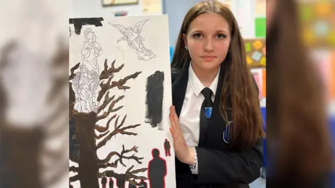 A teenage girl with long brown hair wearing a black school uniform holds up a canvas with a painting of what appears to be an angel on top of a tree.