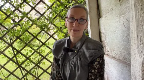 Kelmscott Manor curator Kathy Haslam smiling for the camera. She is sitting inside the manor's summer house. Greenery could be seen growing around the fence behind her.