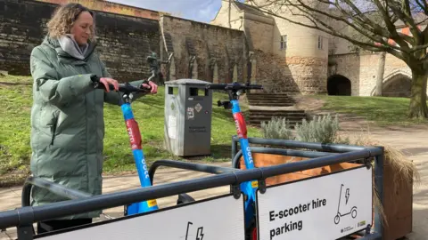 A woman is preparing to try out one of two blue electric scooters which are parked in an official parking place for scooters outside Nottingham Castle