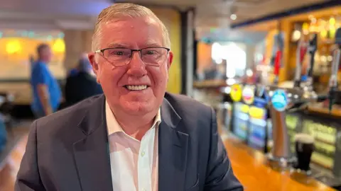 Andy Kerr, who has a short grey hair and glasses, smiles while leading on a wooden bar. He is wearing a business suit.