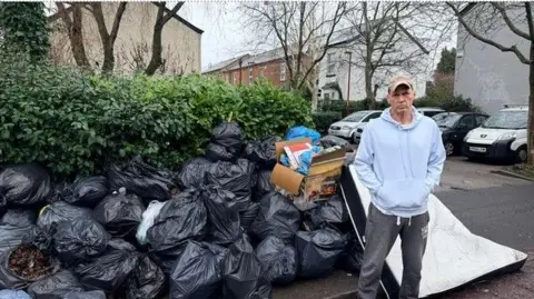Gabriel Bononi A pile of black bin bags stacked up on a street. The rubbish is nearly as tall as a nearby hedge