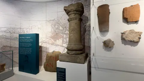 Pieces of stone attached to a white wall on the right. In the middle is a stone column with circular detailing on a white plinth. There is a modern blue plinth to the left with text on.