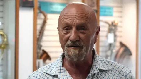 Backhouse stands inside a music shop, speaking to a reporter. He is bald, tanned and wears a checked shirt. He has silver stubble.