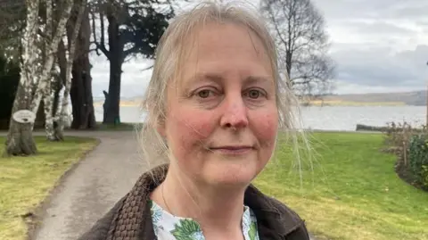 Donna Peacock is standing outside in a green space with trees and the waters of the Beauly Firth beyond. She has fair hair which is tied up and is wearing a brown jacket over a blouse with a floral print.
