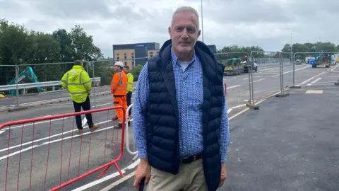 A man with light grey hair wearing a blue gingham shirt and a blue gilet is standing in front of a bridge which has been cordoned off with railings. Engineers wearing high-vis clothing are standing on the bridge, there is an excavator in the background and a number of other vehicles.