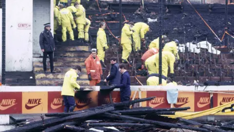 A stand at Valley Parade in the aftermath of the fire. Wooden posts are on the ground burned while people in hi-vis jackets inspect the scene.