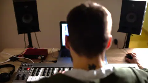 Stock image of a rear view of a musician sitting at a desk in front of a keyboard using his computer