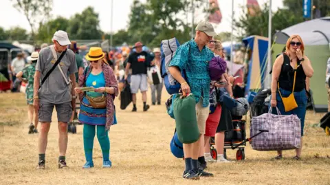 Garry Jones Photography A group of people walk onto site carrying camping equipment. Lots of people can be seen doing the same in the background, with tents running down either side of a large path. 