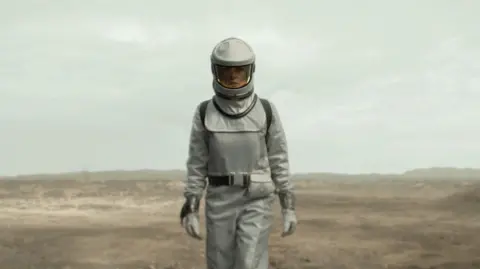 Apple TV+ A woman wearing a protective white suit and helmet with a closed visor stands looking directly at the camera with a crater wasteland behind her.