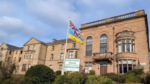 Derbyshire Dales District Council An image of a building with a sign that says DERBYSHIRE DALES DISTRICT COUNCIL. There is also a Union Jack flag and a Ukraine flag on a flagpole.