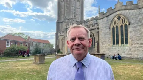 Church warden David Smith stands in the church grounds, with the church in the background. He wears a blue shirt and blue tie. Two people can be seen sitting against the church wall in the background.