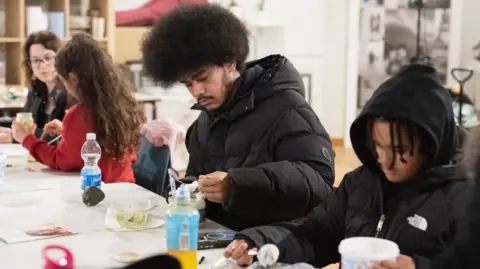 Marnie Bee Photography A woman with long hair and a young girl with long curly hair and a man and a younger boy with afro hair are sitting at a long table and adding ingredients to a hair mask. 