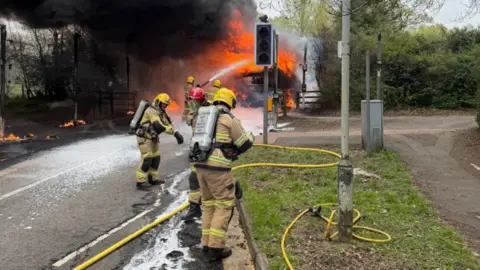 Shropshire Fire and Rescue Service Five firefighters are wearing helmets and brown clothing. Yellow hoses are on the ground in a street. A set of traffic lights is in the middle of the photo and a large fire is in the background.
