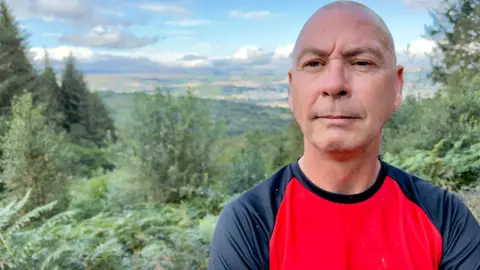 Colin, wearing a scarlet long-sleeved shirt with navy arms and looking at the camera. In the backdrop is a mass of green forest, and a broad sweep of countryside under a summer sky.