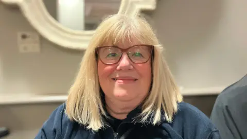 A woman with short blonde hair wearing glasses and smiling, whilst wearing a blue fleece. 