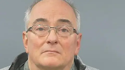 Mugshot of an older man in glasses and a grey jacket