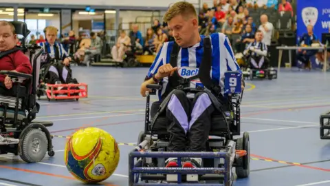 The Albion Foundation Powerchair Football player coach Chris Gordon in action. He is wearing his Albion kit of white and blue and there is a football in front of his chair. We can see spectators behind him cheering the players on. There are some other players behind Mr Gordon.