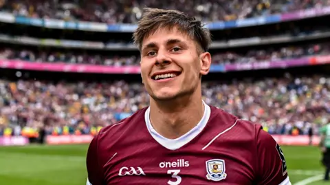 Getty Images Seán Fitzgerald is standing in a pitch in the middle of a huge stadium. He's wearing a maroon GAA jersey. The stadium is full of people. 