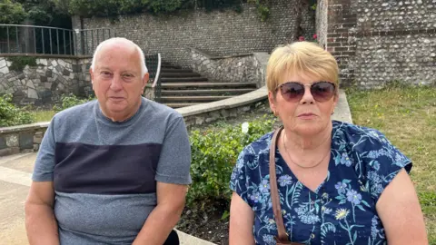 George Carden/BBC Colin and Gillian sat on a paved wall near a bus stop where the shelter has been removed