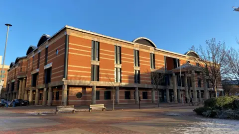 A large three storey court building made from red bricks, with long narrow dark windows and a pyramid shaped porch roof supported by four large stone columns around the main door.