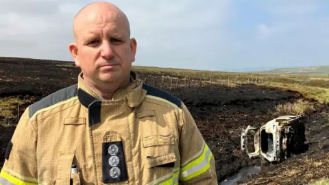 BBC/Victoria Scheer Poynter at the scene of the fire. He is wearing tan firefighting protective clothing with fluorescent yellow and reflective safety strips on the sleeves and torso. Several insignia are visible on the front of the jacket, along with a pen or small tool tucked into a chest pocket.

Behind him, a large area of vegetation has been burned, leaving the ground blackened and charred. A fence line runs across the landscape, with wooden fence posts stretching into the distance. The scorched terrain extends across much of the left side of the image and into the background.

On the right side of the image, a severely burned vehicle lies on its side in a drainage ditch. The vehicle's exterior has been almost completely destroyed by fire, exposing much of the metal framework. The ditch appears to contain a small amount of water, and the surrounding grass and vegetation show signs of fire damage.

The setting is an open countryside area with rolling hills visible on the horizon. 