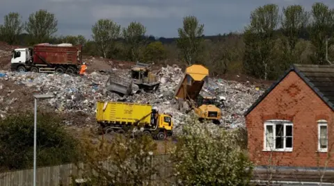 Four lorries working on a large pile of landfill waste immediately behind a modern, brick-built house.