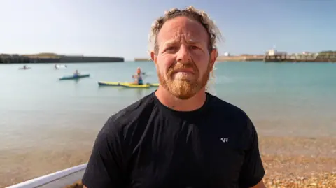 Tim Everest/BBC Wesley Horscroft pictured on Kingston Beach, Shoreham. He has blondish hair and a ginger beard. He is wearing a black t-shirt. Paddleboarders can be seen in the background.