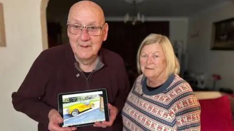 Frank Dowsett is wearing a dark red jumper and is holding an iPad with a picture of his bright yellow Morgan classic sports car. His wife Millie is wearing a patterned jumper next to him. They are both standing in their lounge