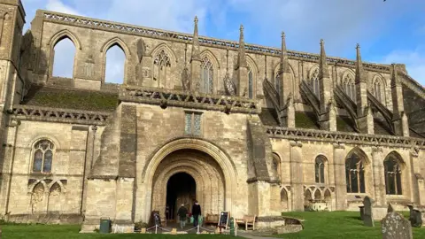 A historic abbey with graveyard outside under a blue sky. Tall with large archways and windows.