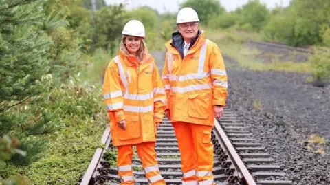 A blonde woman standing next to an older man. Both are wearing orange hi-vis jackets and trousers and hard hats. They're standing on a railway line surrounded by green bushes.
