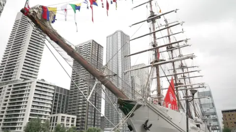 Modern skyscrapers and a grey sky in the background, the bow of a tallship with sails folded away in the foreground. The hull is grey, the masts are wooden and there are colourful flags from different nations haging from her rigging
