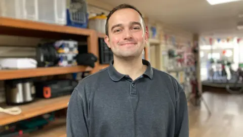 Frank Kibble wearing a dark grey polo shirt and smiling at the camera. He is standing in the storage unit of Stroud's Library of Things in front of a large wooden shelving unit. Frank has short brown hair, blue-grey eyes and is clean shaven.