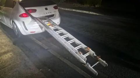 A white Seat Leon stopped on the side of the road, it has a large set of ladders sticking out the boot which is partially open.