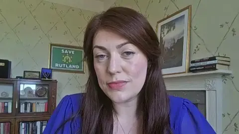 A head and shoulders shot of Alicia Kearns. She has long, brown hair and is wearing a blue blouse. Behind her is a brown cabinet with books in, a green, framed print reading "SAVE RUTLAND", as well as prints and books stacked on a fireplace.