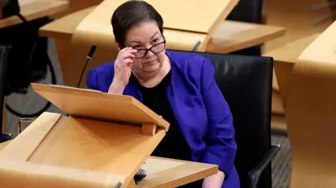 Getty Images Jackie Baillie seated at a wooden desk in a parliamentary-style chamber, adjusting a pair of glasses. A microphone, black chair and surrounding wooden desks are visible.