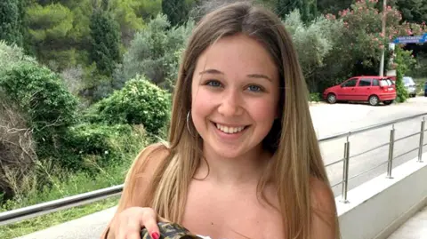 Ellie Gould has long hair and is pictured on a balcony, smiling at the camera. There is a red car in the background. 