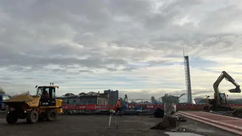 The building site for the new park. There are two diggers and workmen on the site. The pump house, the Science Centre and bridges over the Clyde can be seen in the background. 