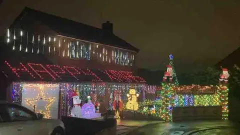 A house covered in Christmas lights with the words "Happy Xmas" projected onto a sloping roof. There are lit-up figures of Father Christmas and snowmen at the front.