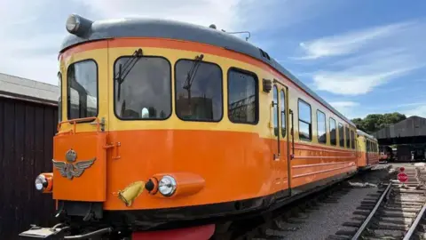 NENE VALLEY RAILWAY A orange rail cart, made up of two carriages, is on the tracks on a sunny day. It is rounded at the sides with yellow paint around the windows, orange on the body and black on the top and bottom.